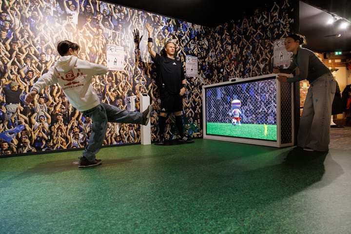 Un garçon fait un tir de foot vers un écran avec un gardien virtuel, tandis que deux adultes l'observent dans une salle avec un mur décoré d'une foule applaudissante.