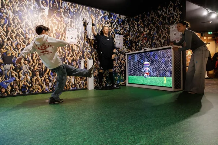 Un garçon fait un tir de foot vers un écran avec un gardien virtuel, tandis que deux adultes l'observent dans une salle avec un mur décoré d'une foule applaudissante.