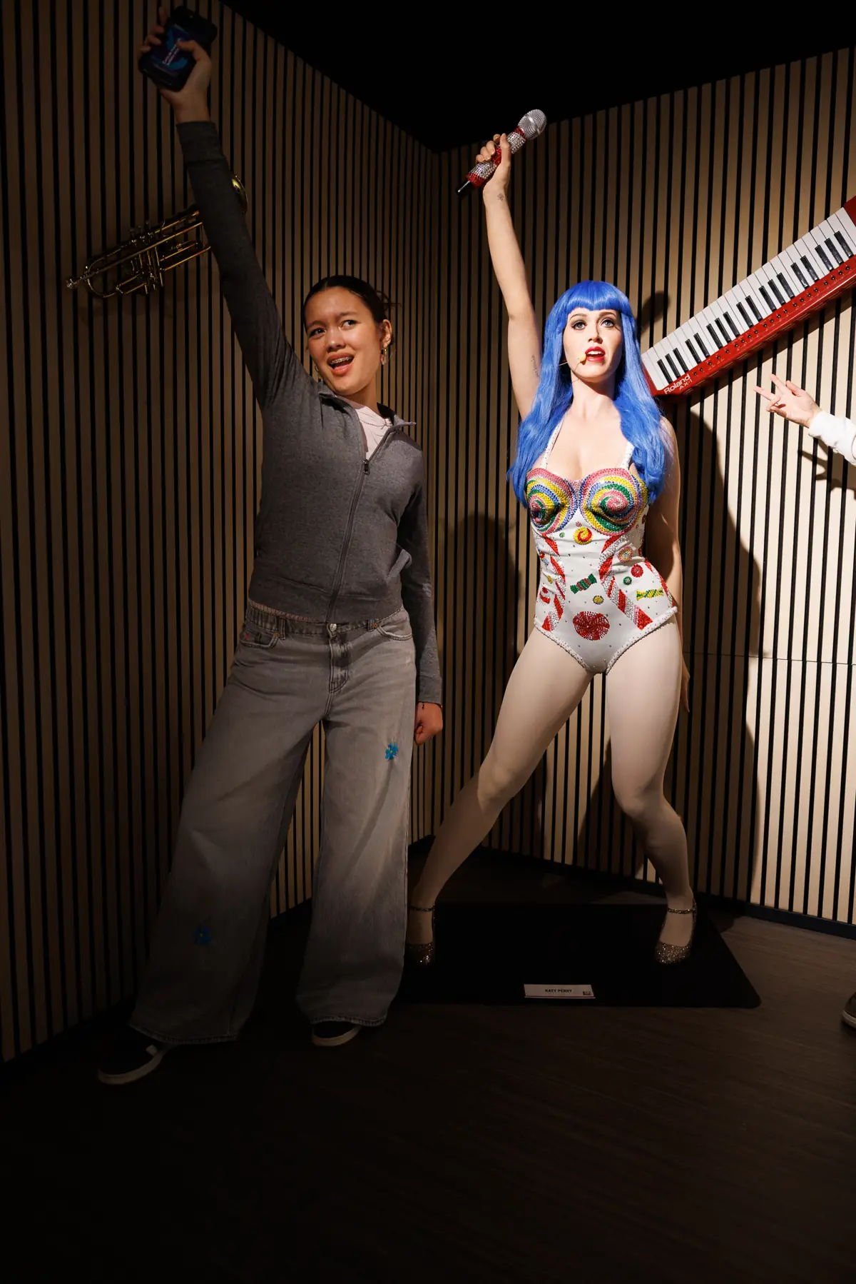 Visiteuse posant bras levé aux côtés de la statue de cire de Katy Perry, perruque bleue et micro, au Wax of Legends de Colmar