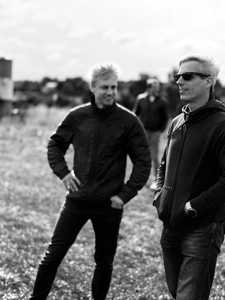 Two men outdoors, one wearing sunglasses and a hoodie, the other wearing a jacket and smiling.