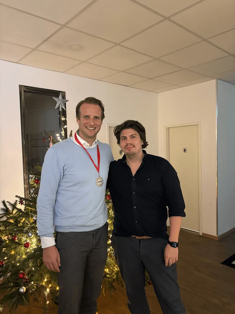 Two men stand in front of a tree decorated for Christmas and smile at the camera.