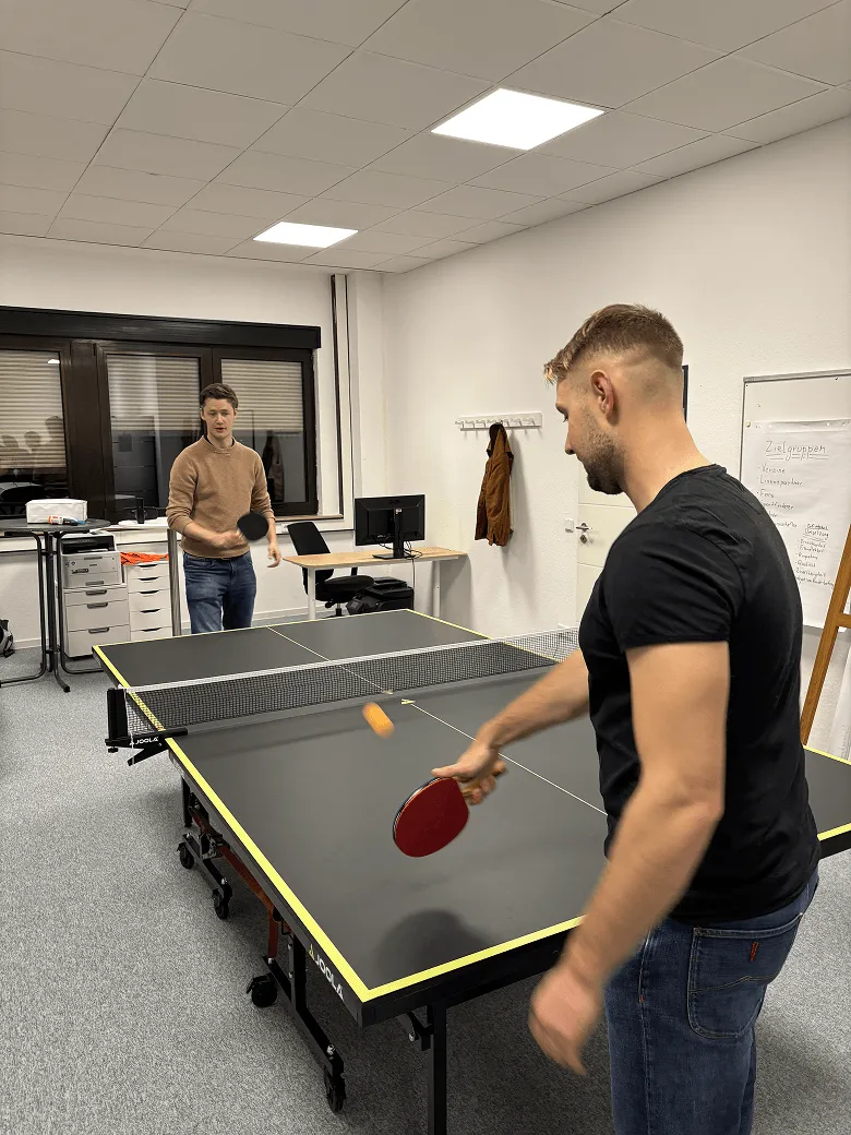 Two men play table tennis in an office with a computer desk and jacket on the wall.