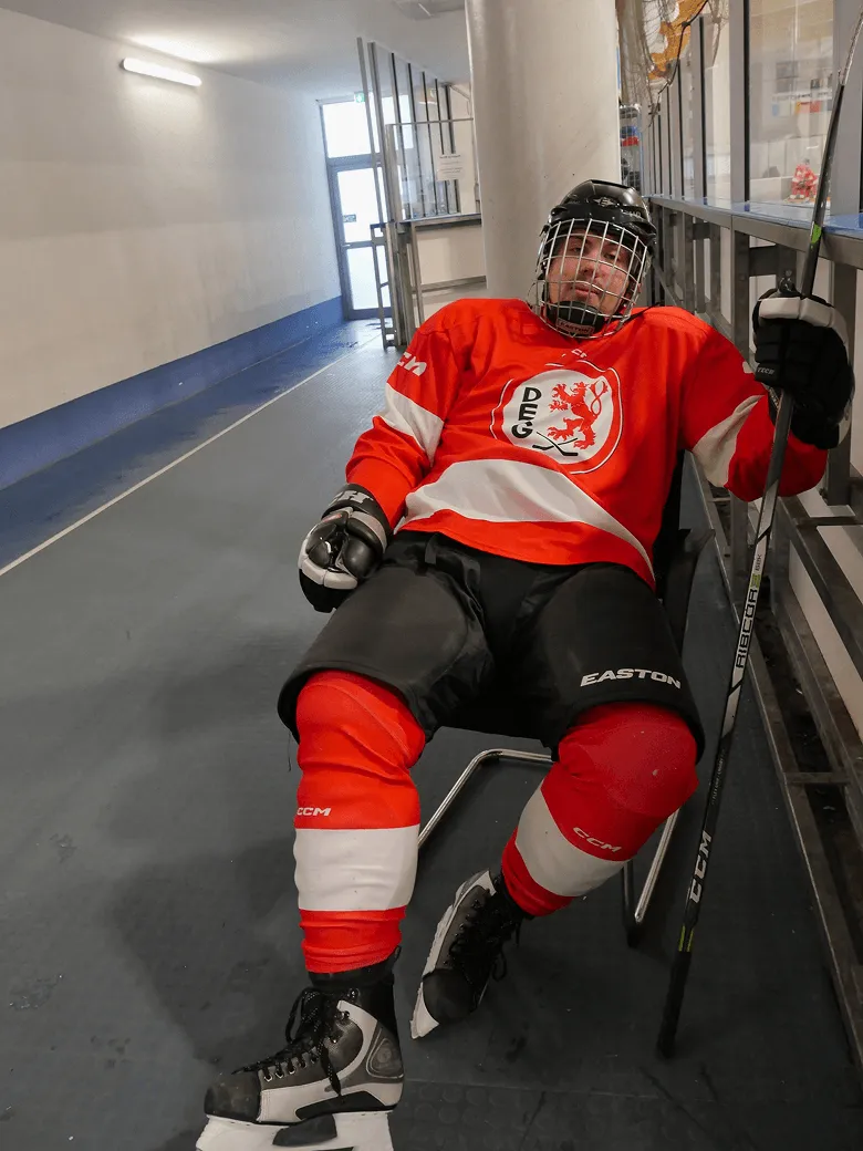 Ice hockey player is sitting in the cabin hall wearing a red jersey with helmet and racket.