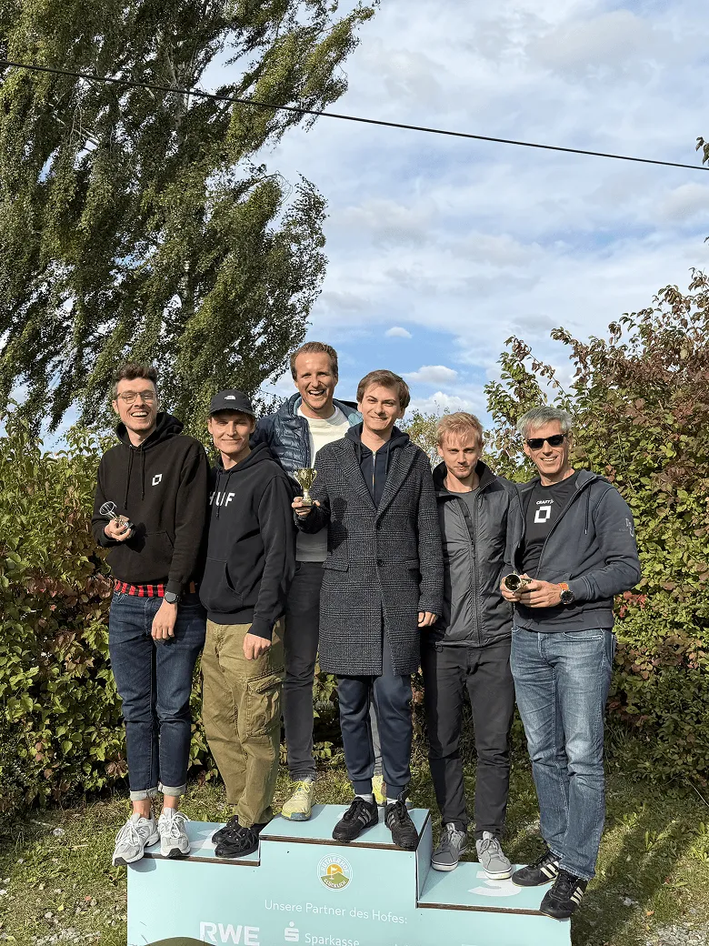 Six men stand smiling on a winners' podium outdoors with trees and sky in the background.