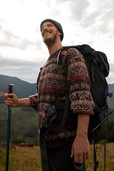 Smiling man wearing a patterned sweater and beanie, hiking with a backpack and trekking pole in a mountainous area.