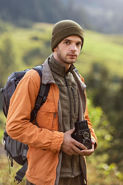 Young man outdoors holding a vintage camera, wearing an orange jacket, olive green beanie, and backpack with a forest background.