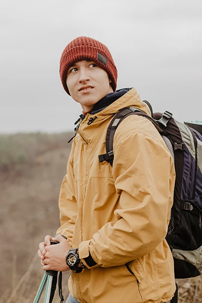 Young man in a yellow jacket and red beanie holding hiking poles with a backpack, standing outdoors on a cloudy day.