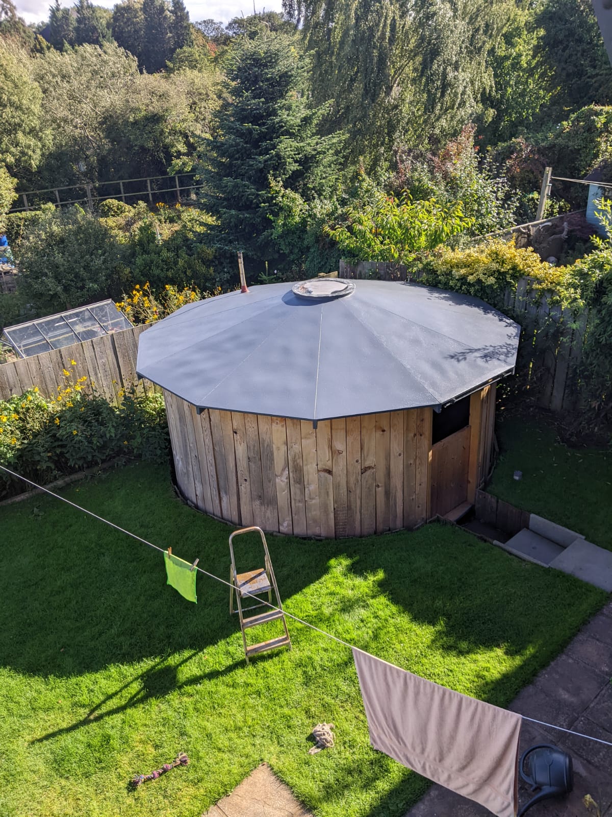 Round wooden garden shed with a dark gray roof in a backyard with green grass, a step ladder, and two towels hanging on a clothesline.