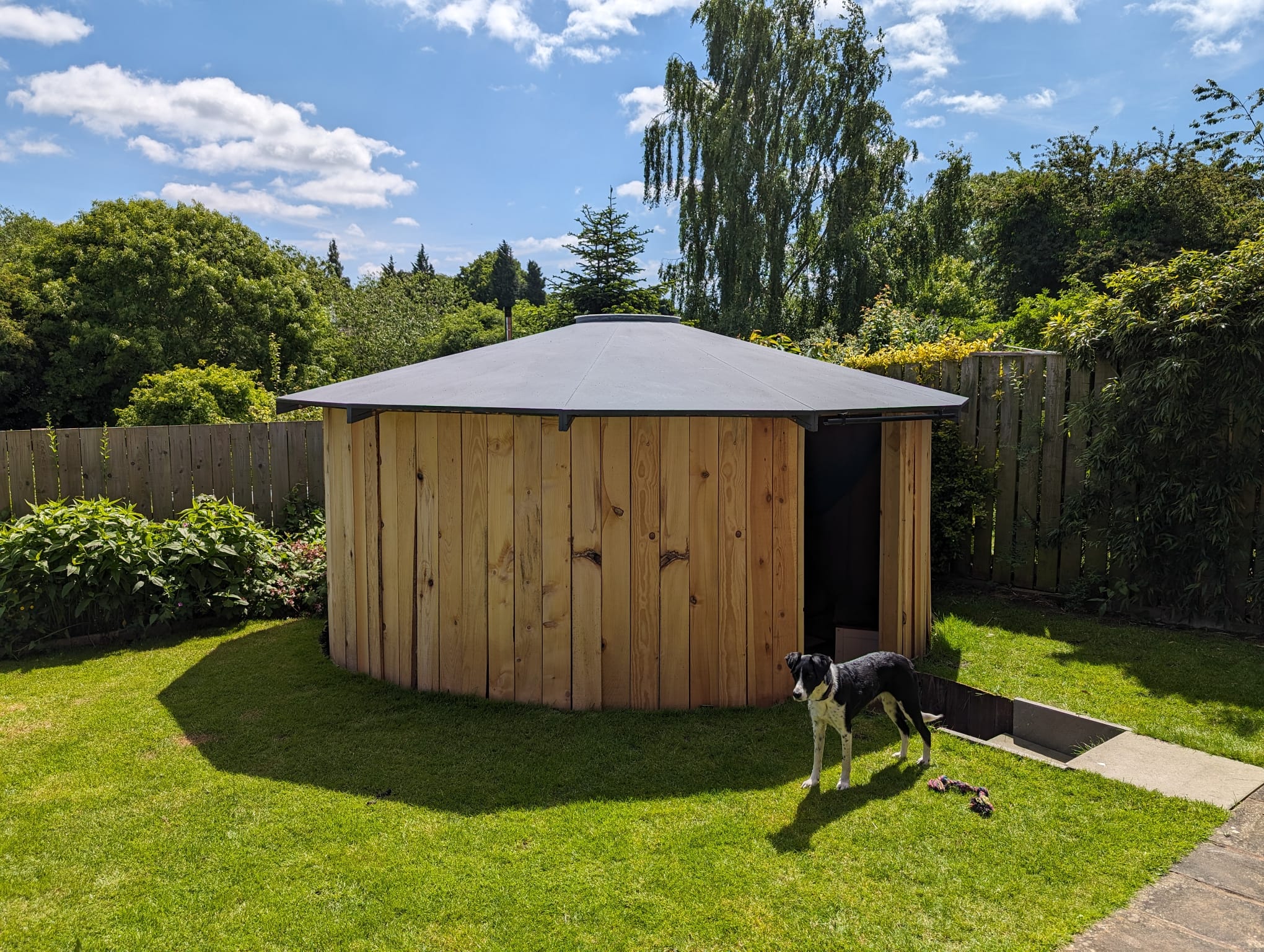 Round wooden garden shed with a gray roof on a lawn with a black and white dog standing beside the open doorway under a partly cloudy blue sky.