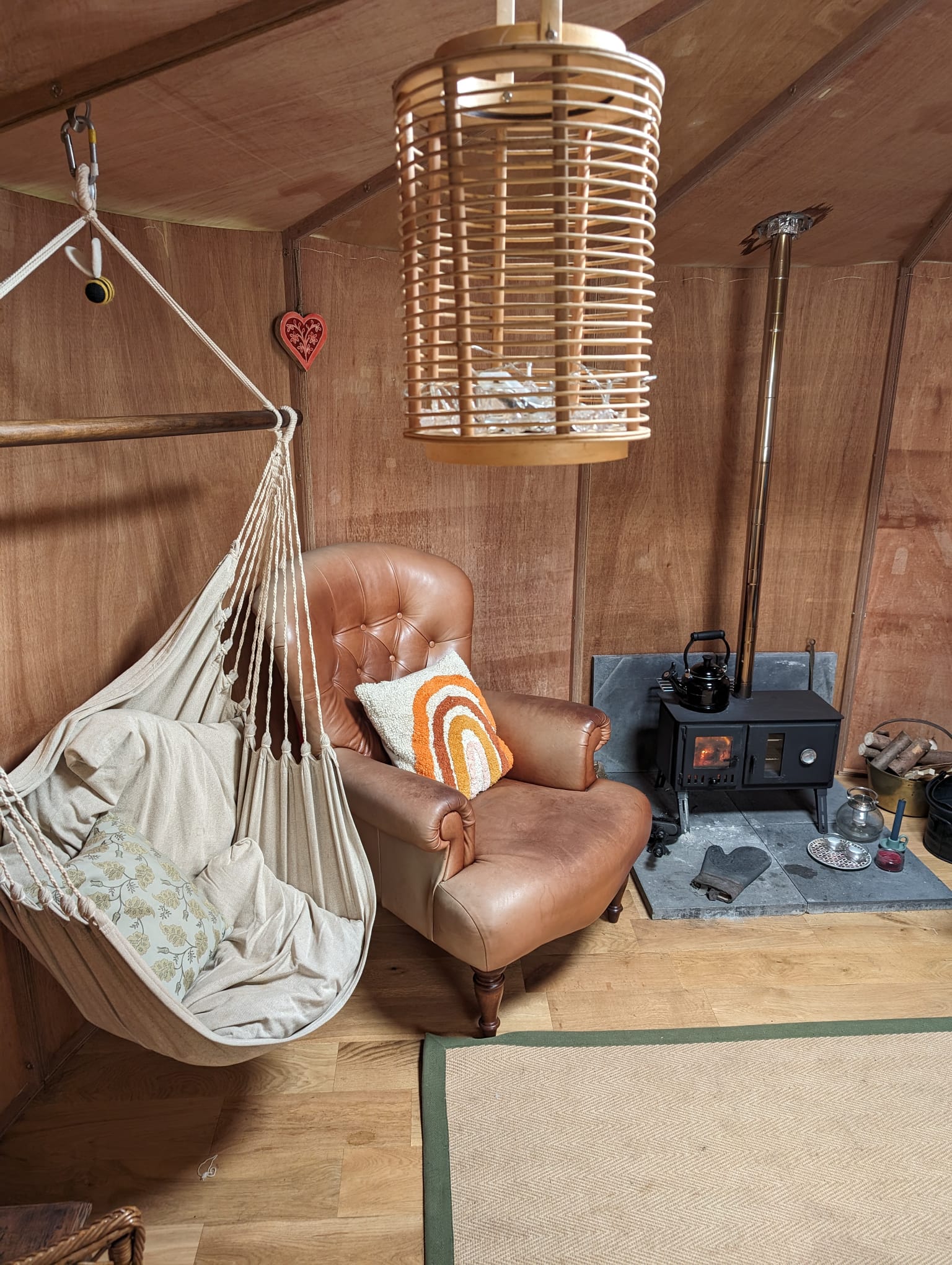 Cozy corner with a leather armchair, hanging fabric hammock chair, and a wood-burning stove with a kettle on top inside a wooden cabin.