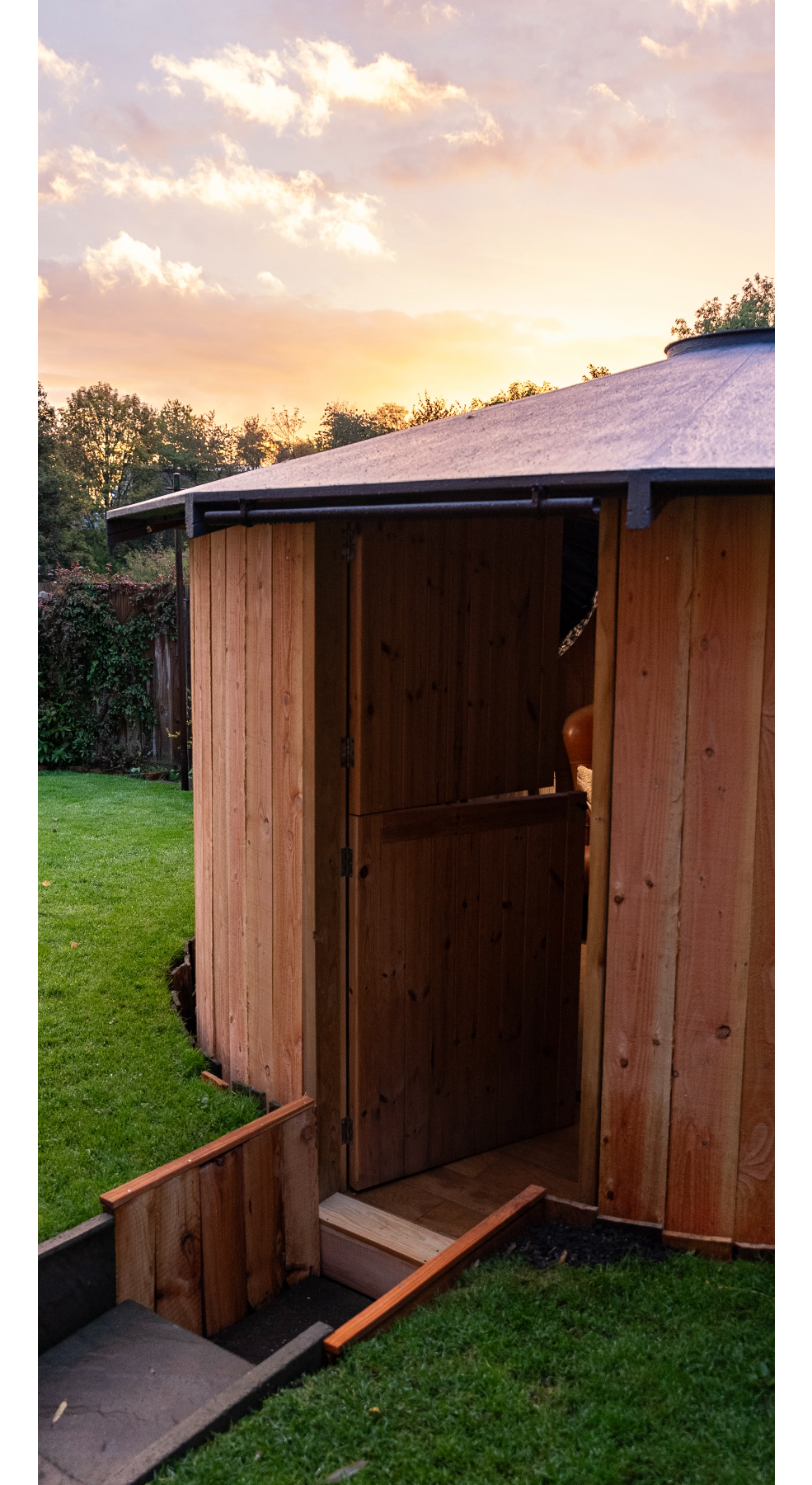 Wooden round garden hut with half-open door at sunset with green grass and trees in the background.