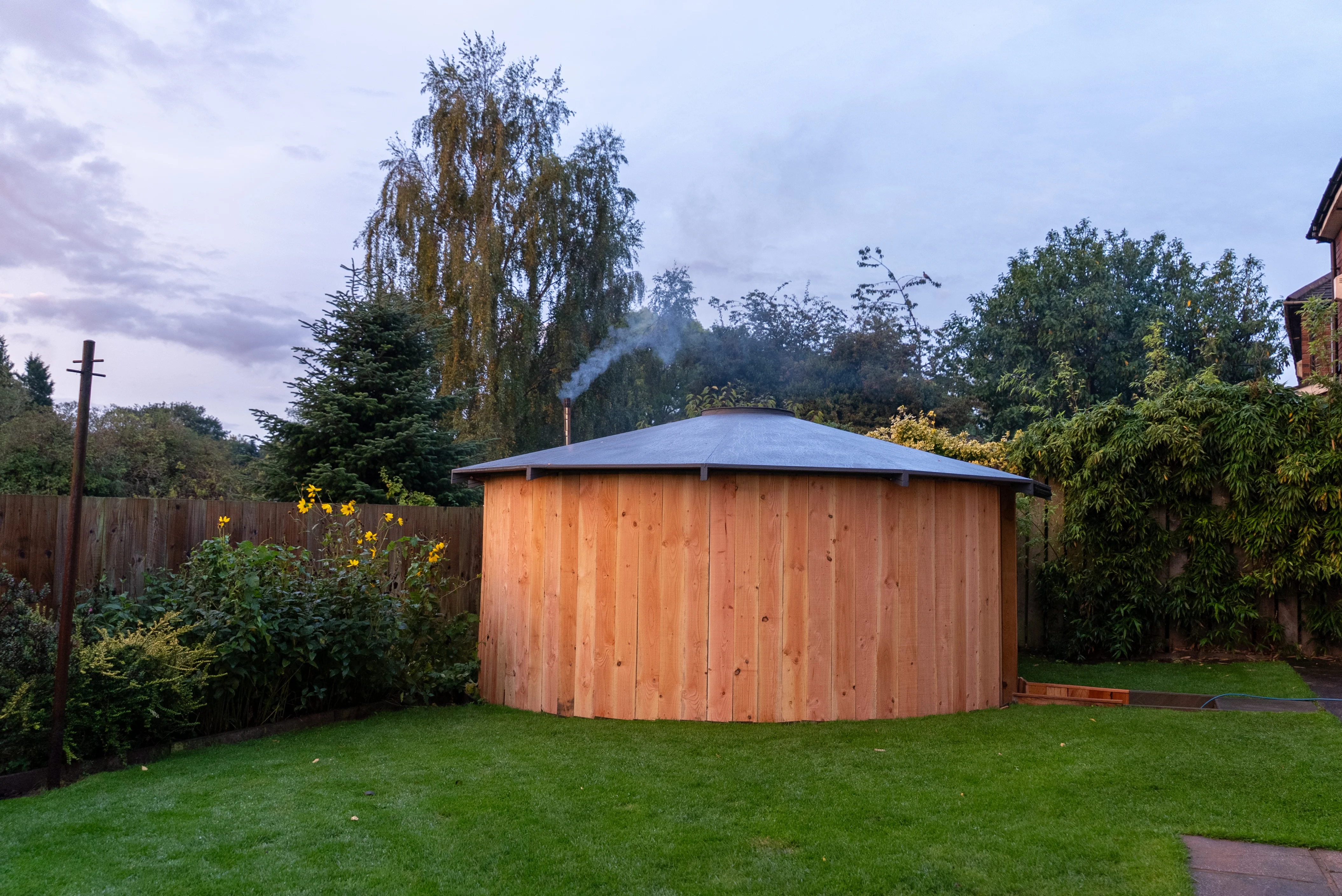 Large round wooden hot tub with smoke coming from a chimney, set on a green lawn in a backyard with trees and plants around.