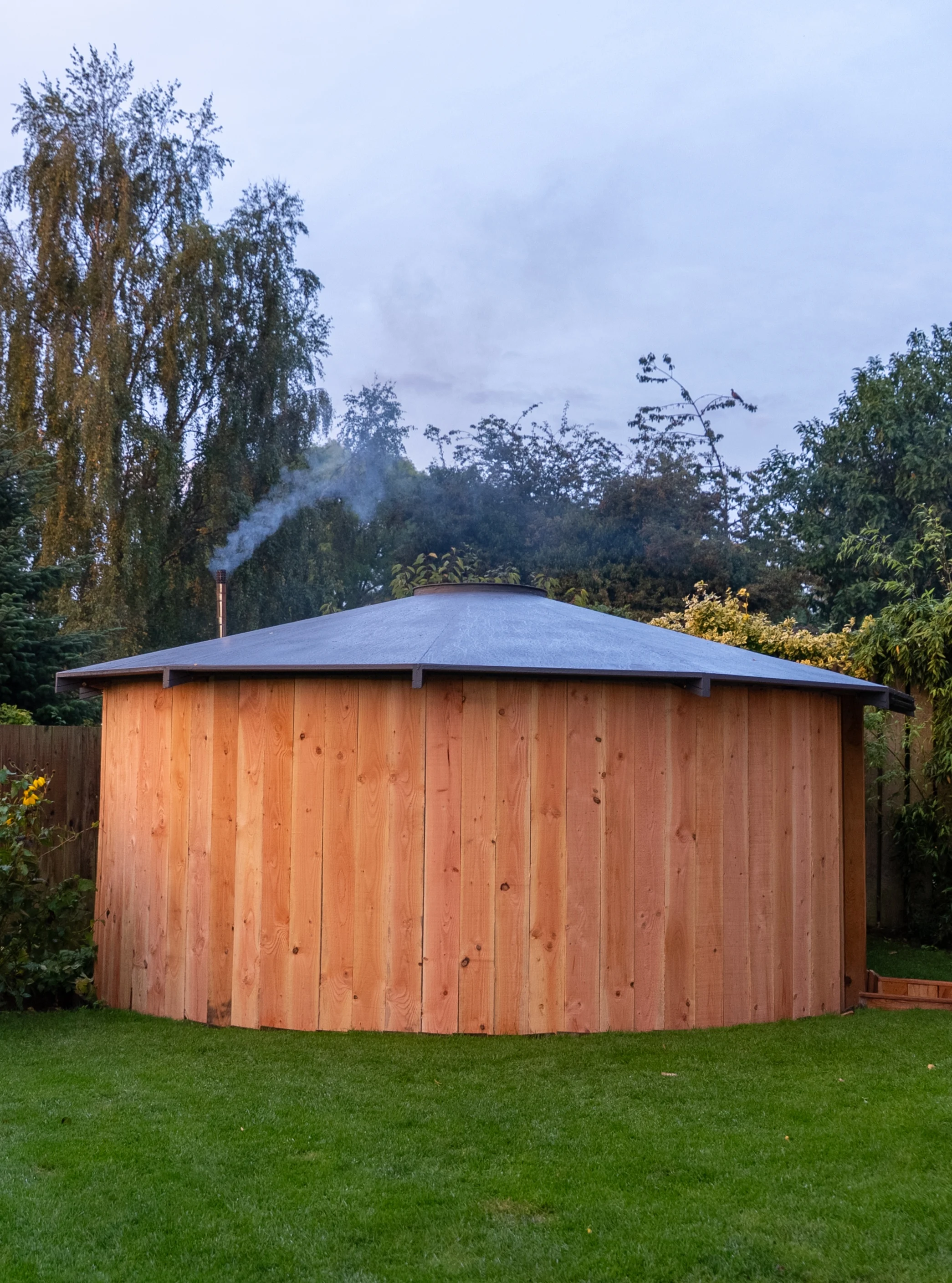 A circular wooden sauna cabin with a conical metal roof and smoke coming from a chimney, situated on green grass with trees in the background.