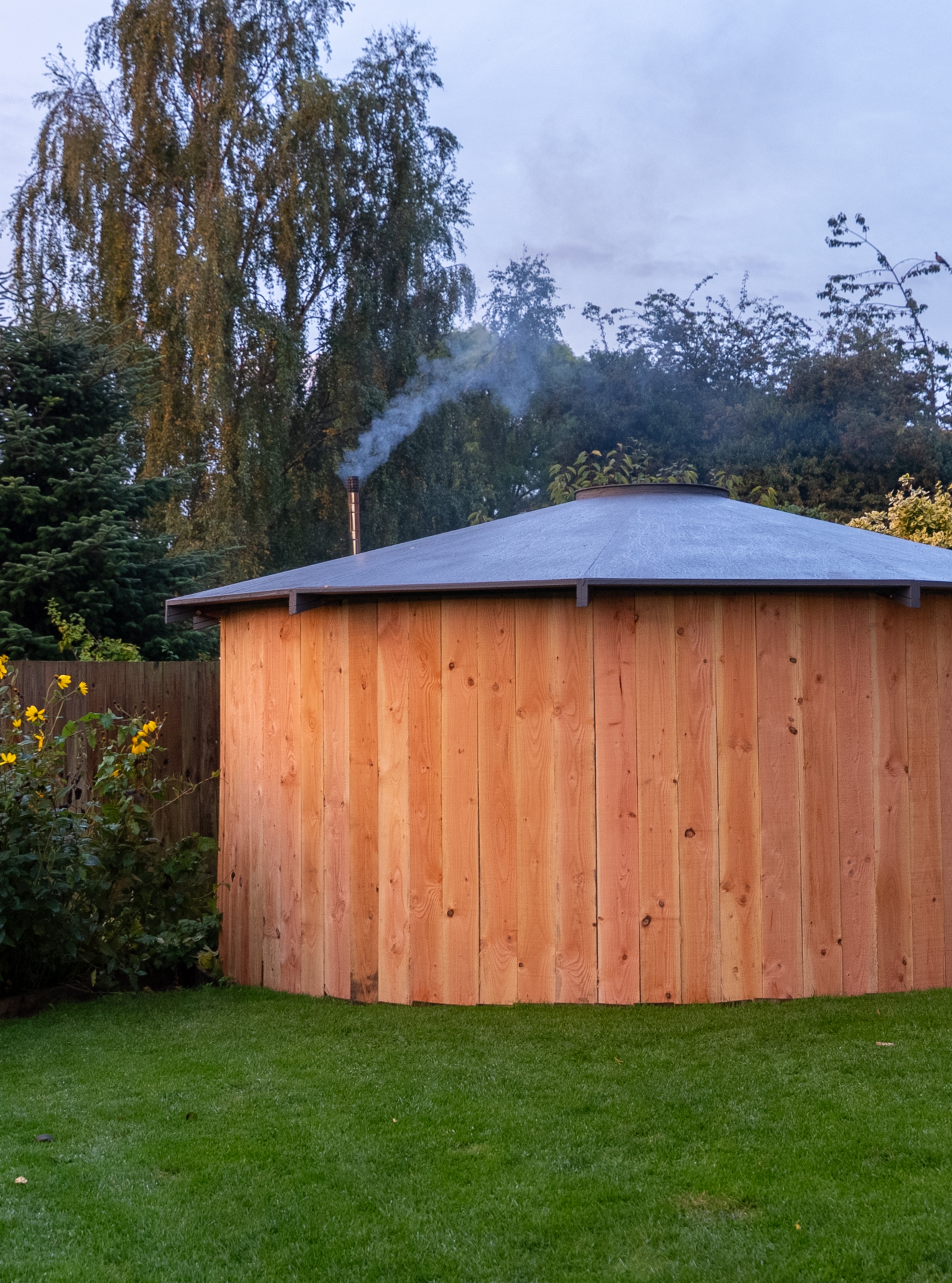 Round wooden outdoor sauna with a metal roof and smoke coming from its chimney in a grassy backyard surrounded by trees and plants.