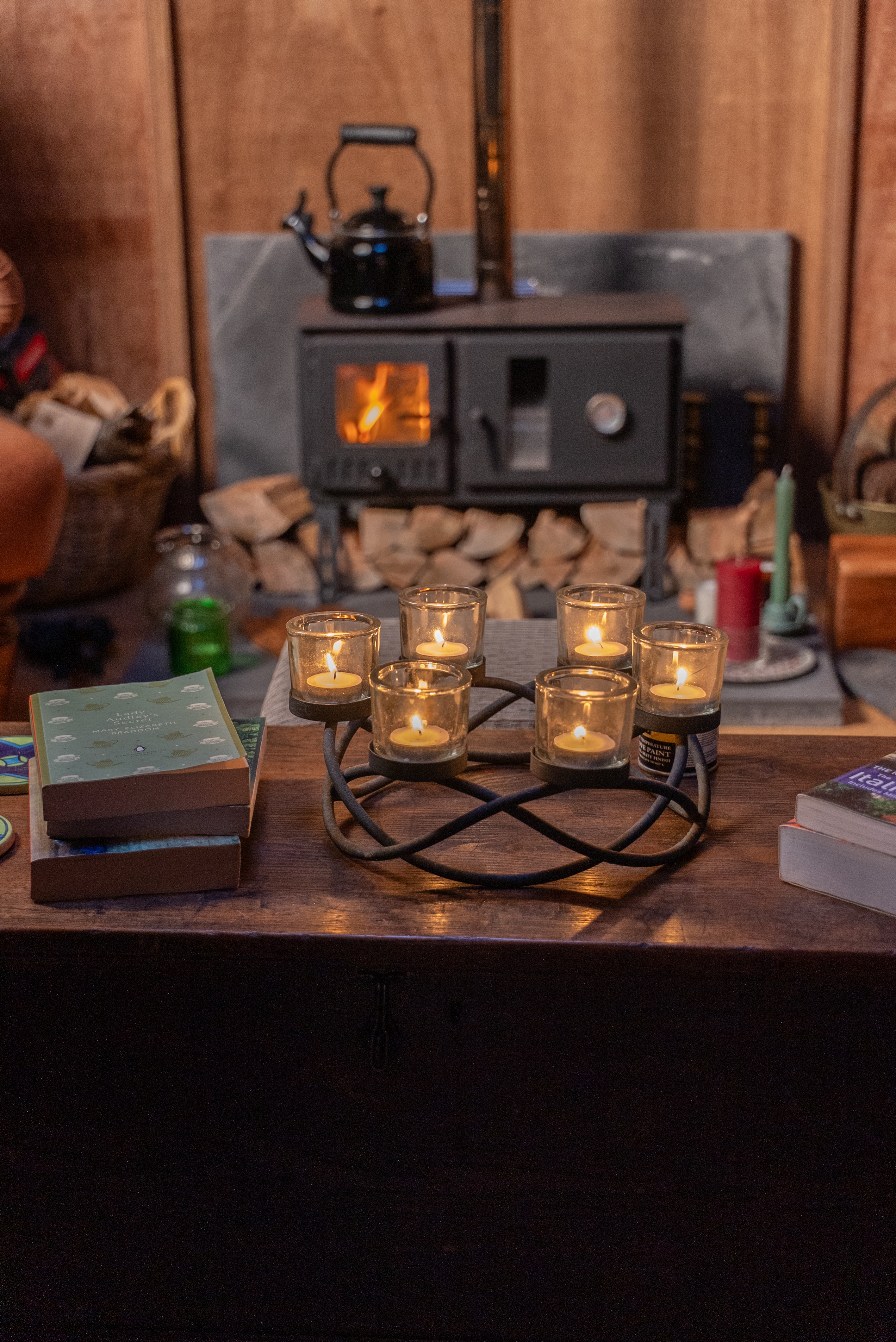 A wooden table with six lit candles in glass holders arranged on a metal wreath centerpiece, stacks of books on either side, and a wood-burning stove with a kettle in the background.