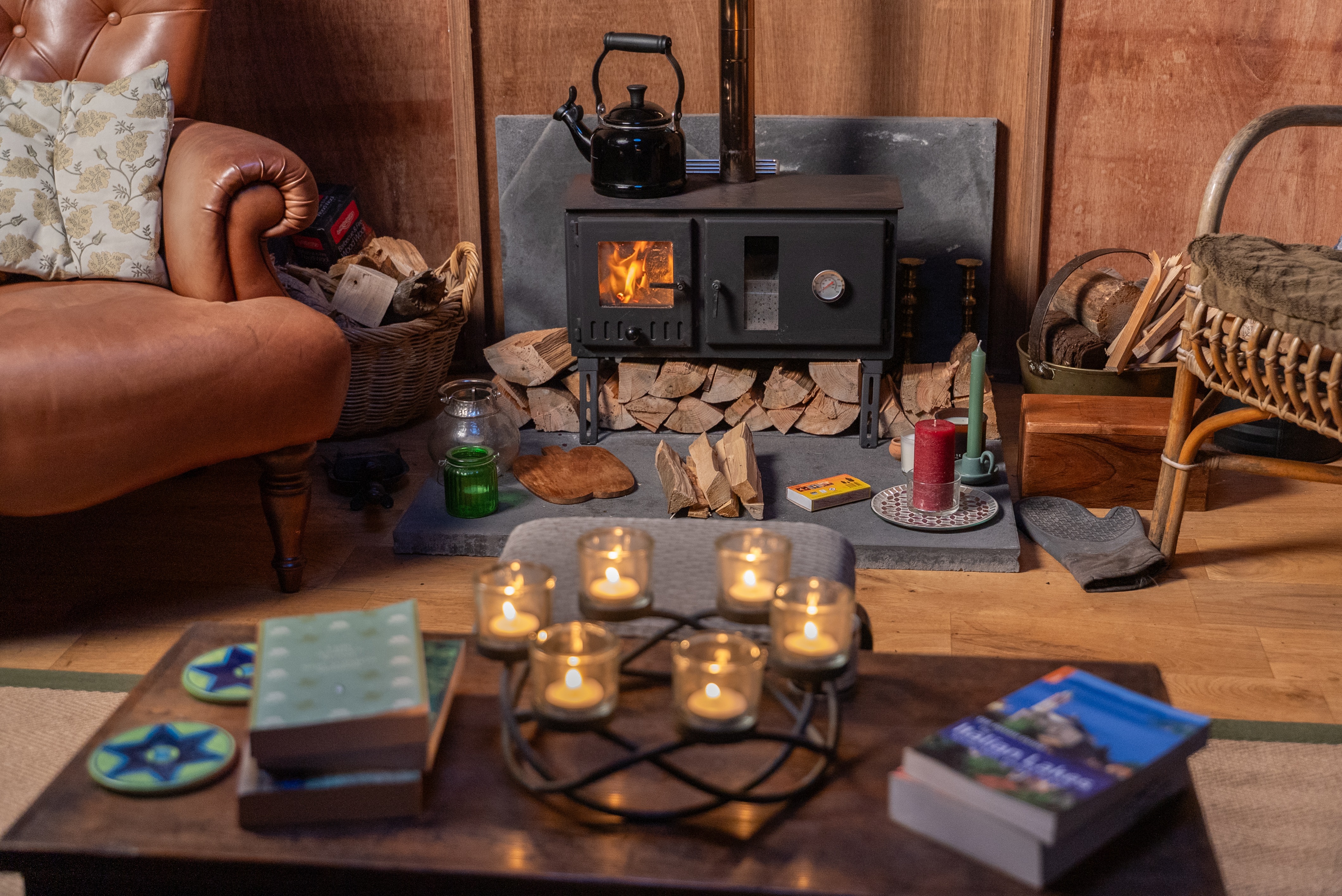 Cozy living room with a lit wood-burning stove, stacked firewood, candles on a wooden table, and a leather armchair with floral pillows.