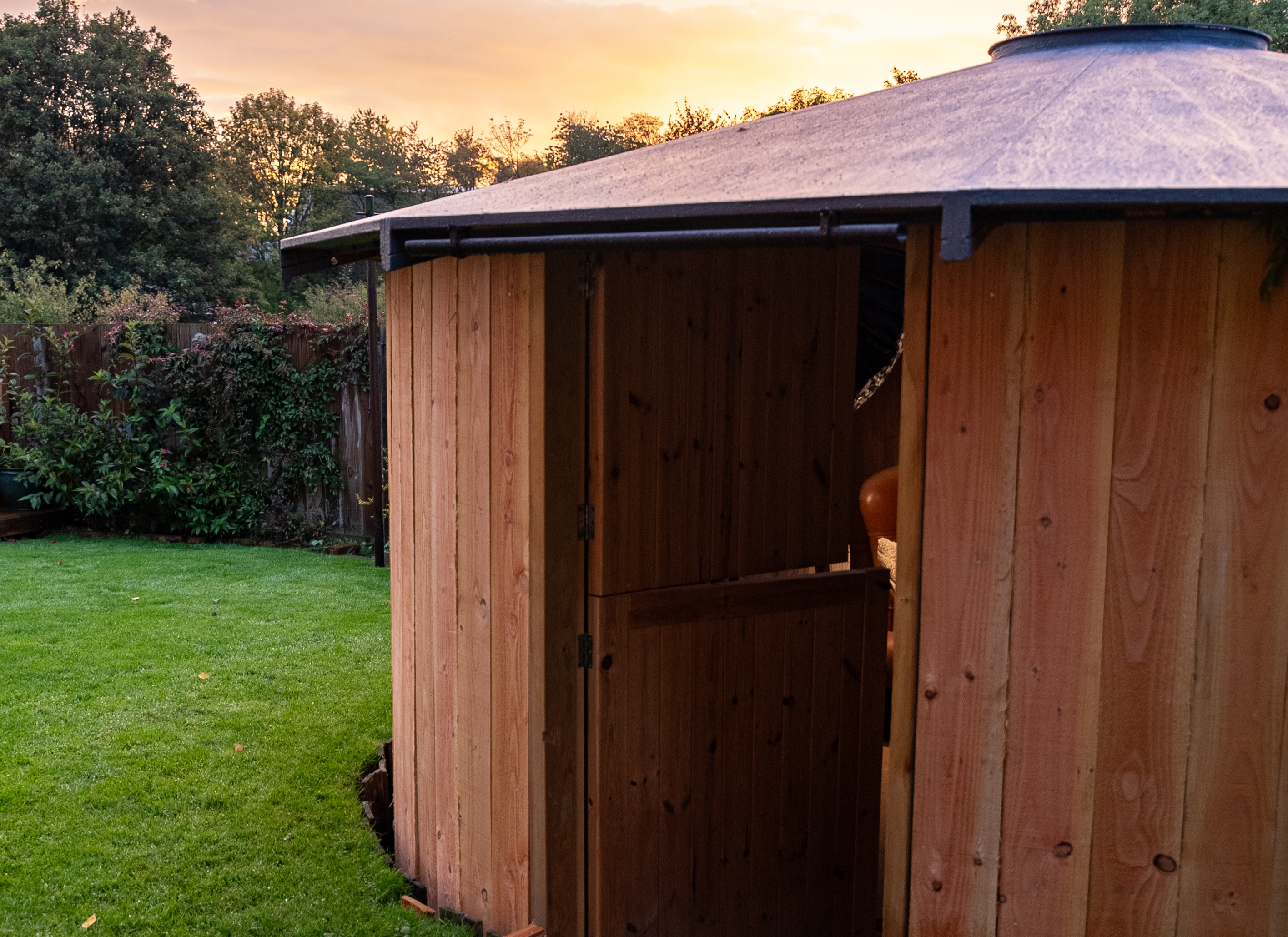 Wooden garden shed with an open door showing a glimpse of a brown chair inside, set on green grass with trees and a wooden fence in the background at sunset.