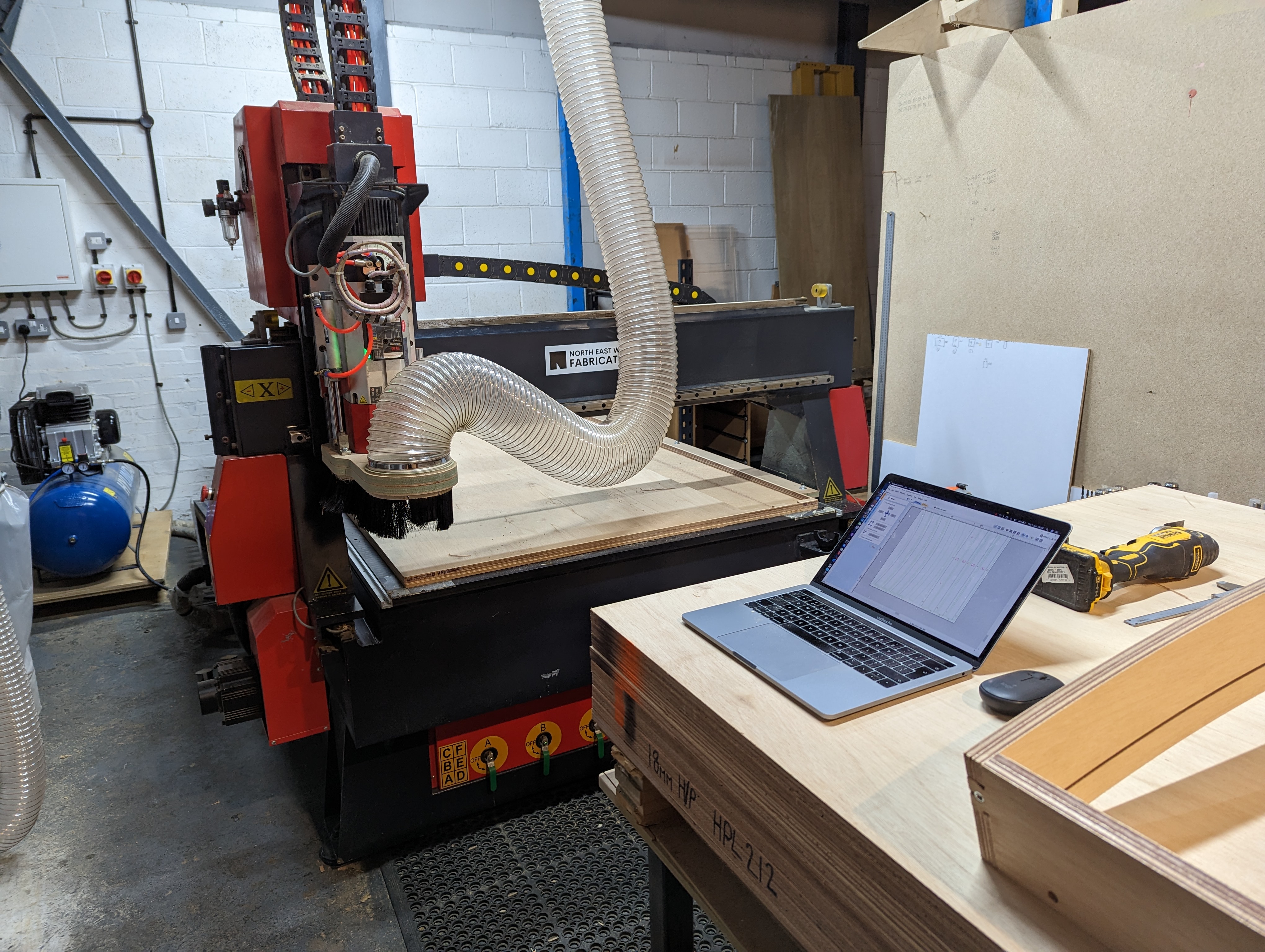 Workshop CNC machine with dust extraction hose, a laptop on a wooden workbench, and power drill nearby.