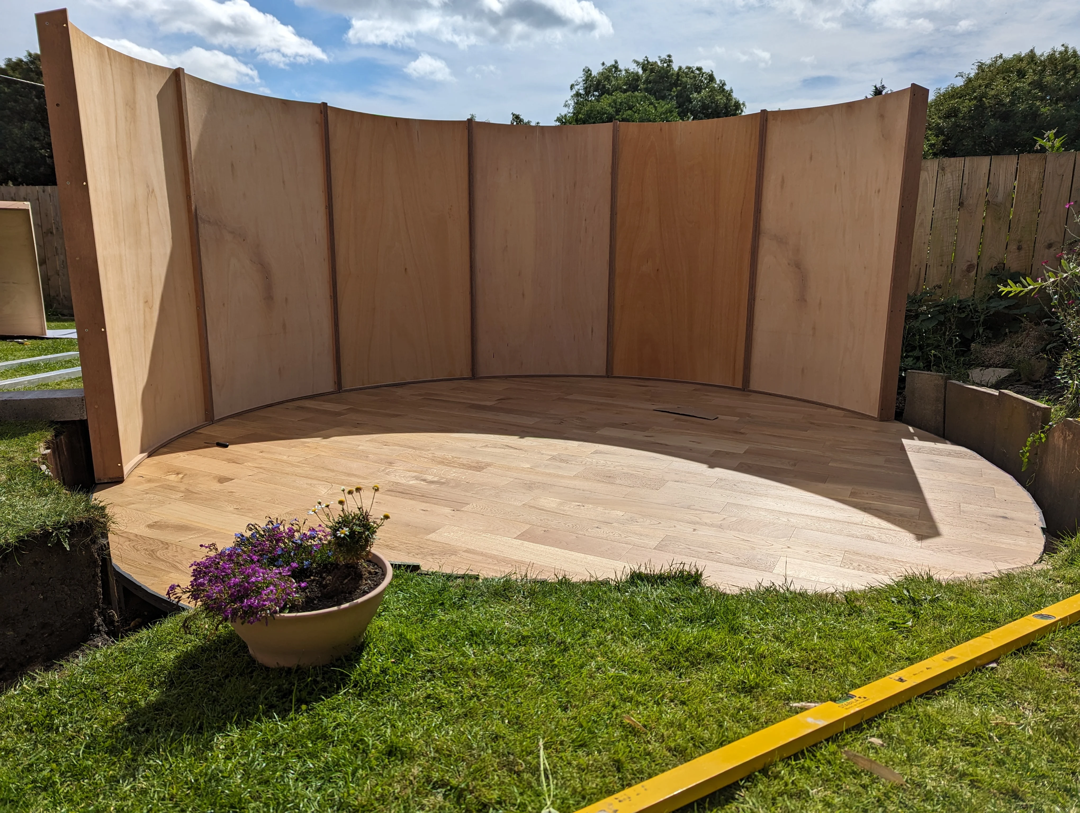 Curved wooden platform with tall wooden panels set up outdoors on grass under a partly cloudy sky.