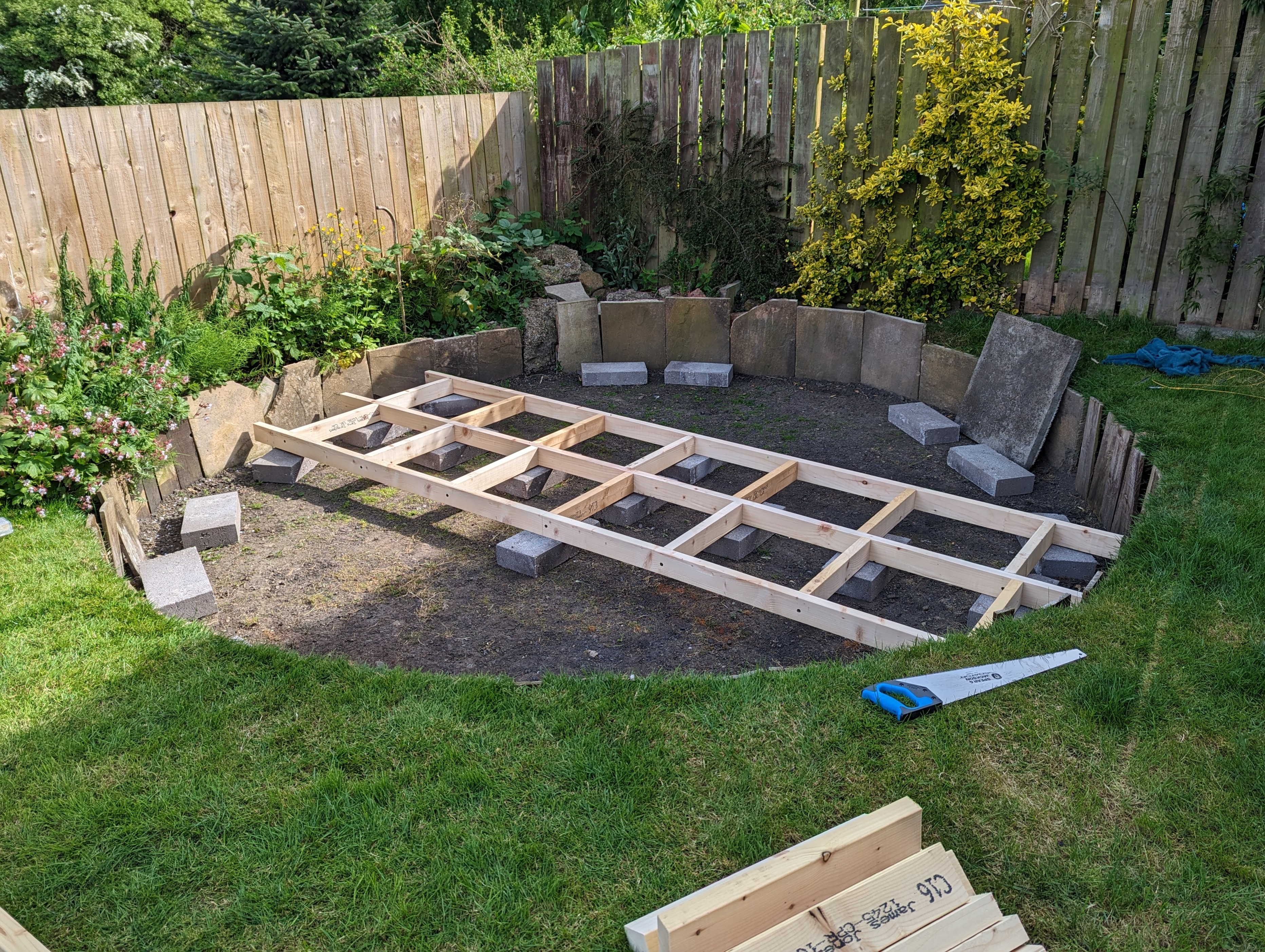 Wooden frame laid out on cinder blocks inside a dug-out circular garden area, surrounded by wooden fence and plants, with a saw on grass.