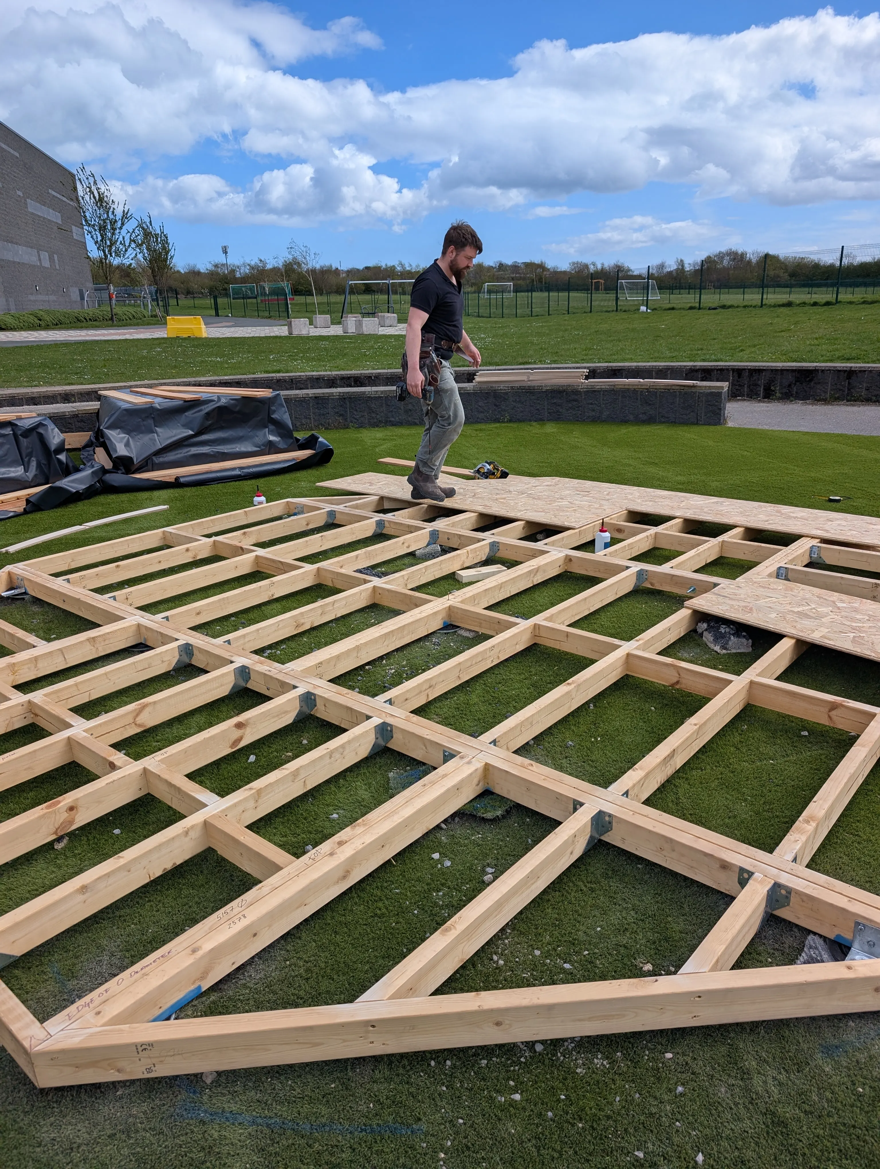 Wooden frame laid out on cinder blocks inside a dug-out circular garden area, surrounded by wooden fence and plants, with a saw on grass.