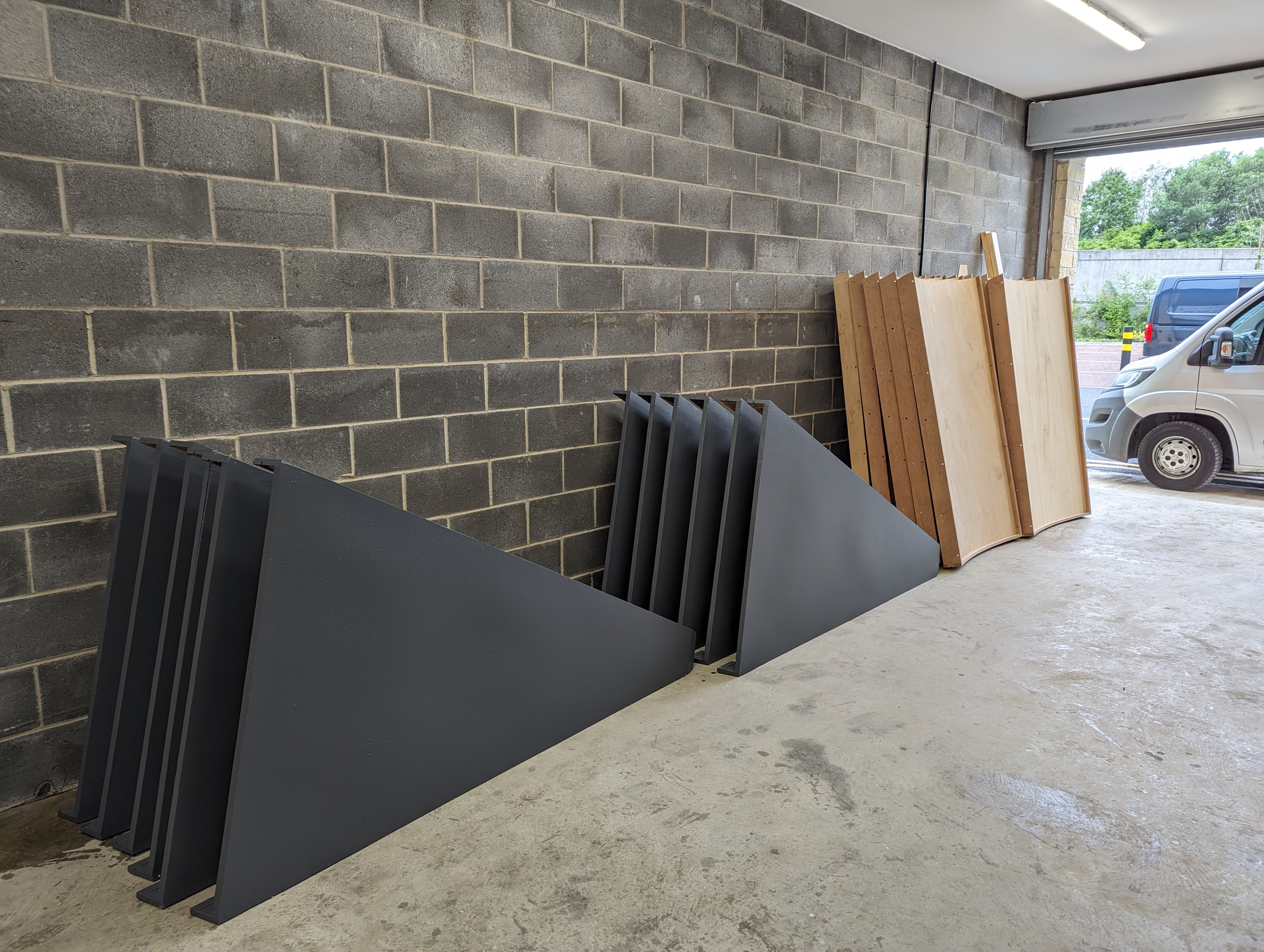 Two stacks of large black triangular metal pieces and several wooden panels leaning against a brick wall inside a garage with a partially visible white van outside.