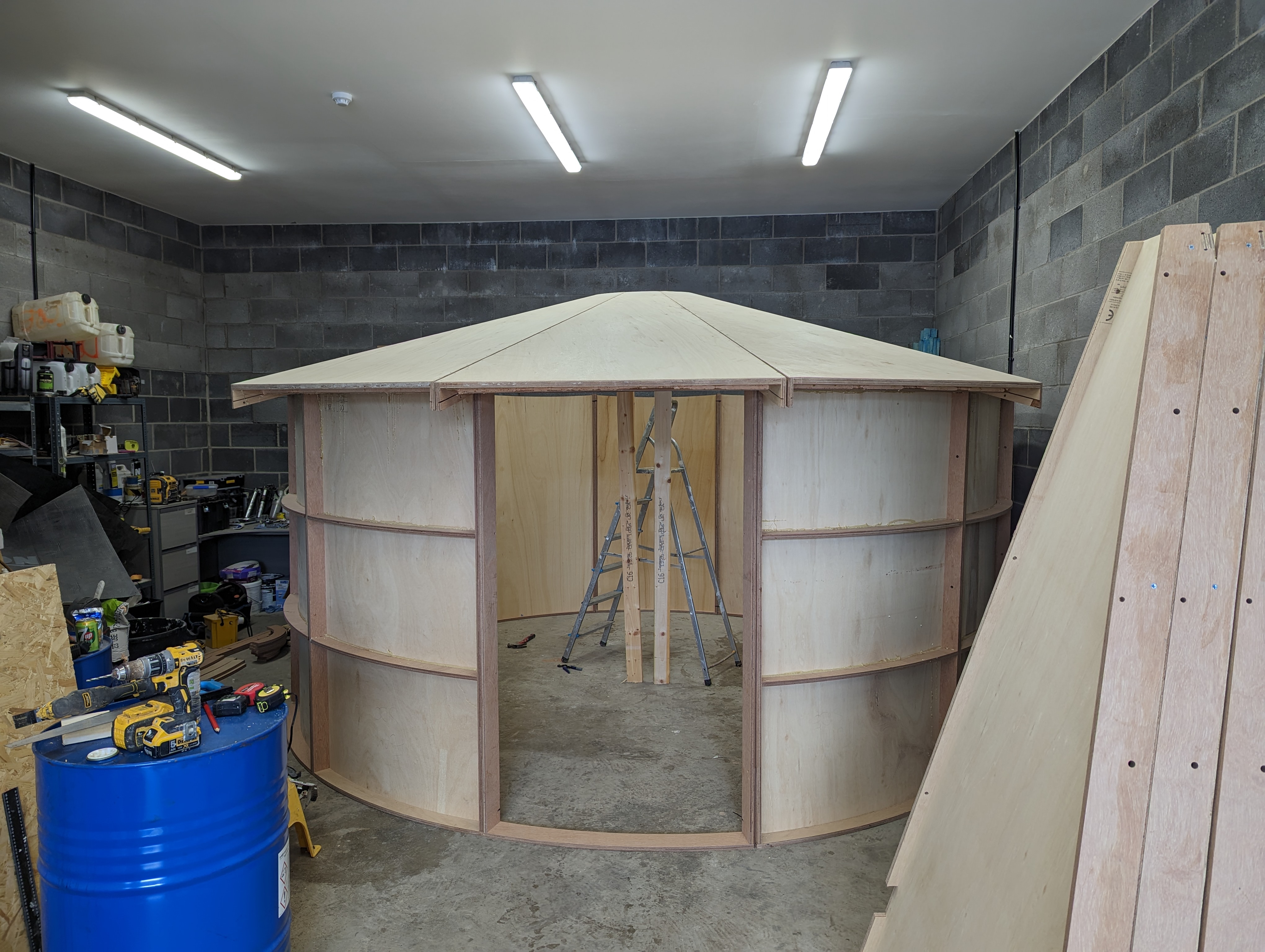 Partially constructed circular wooden structure with an open doorway inside a workshop with tools and ladders.