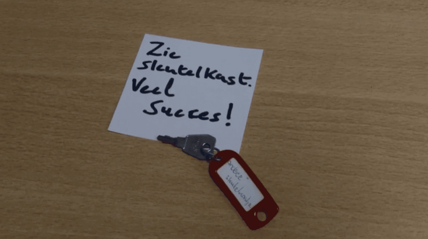 Sleutel met rode sleutelhanger “nieuwe sleutelkast” op hout, naast een briefje: “Zie sleutelkast. Veel succes!”