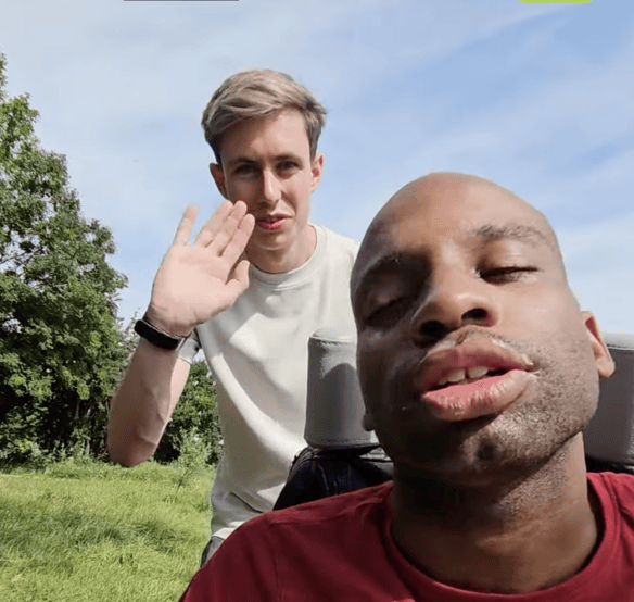 Twee mannen buiten op een grasveld met bomen; één man op de voorgrond in een rood shirt, de ander daarachter zwaaiend in een wit shirt.