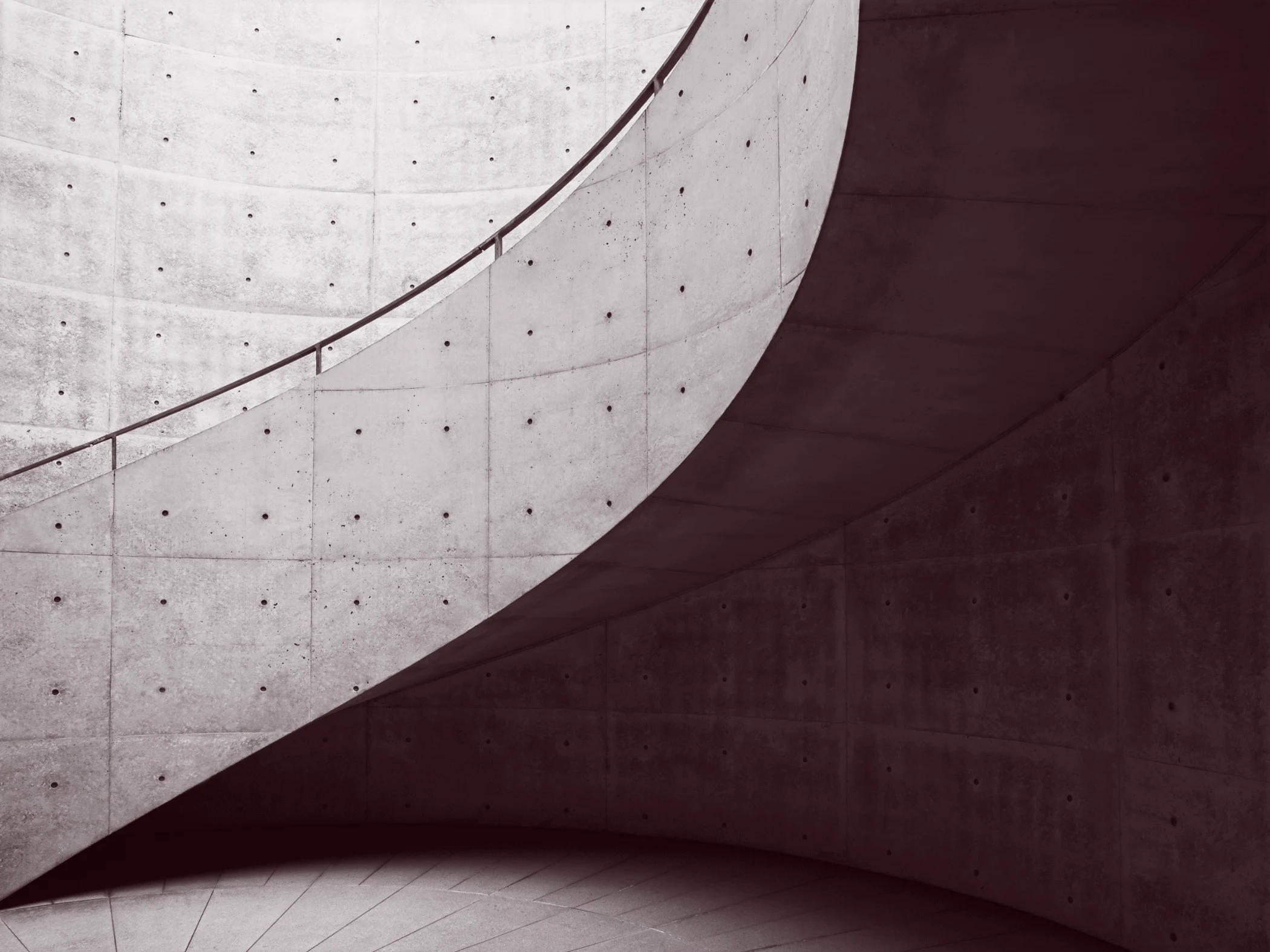 Curved concrete staircase with metal railing casting shadows on a textured concrete wall.
