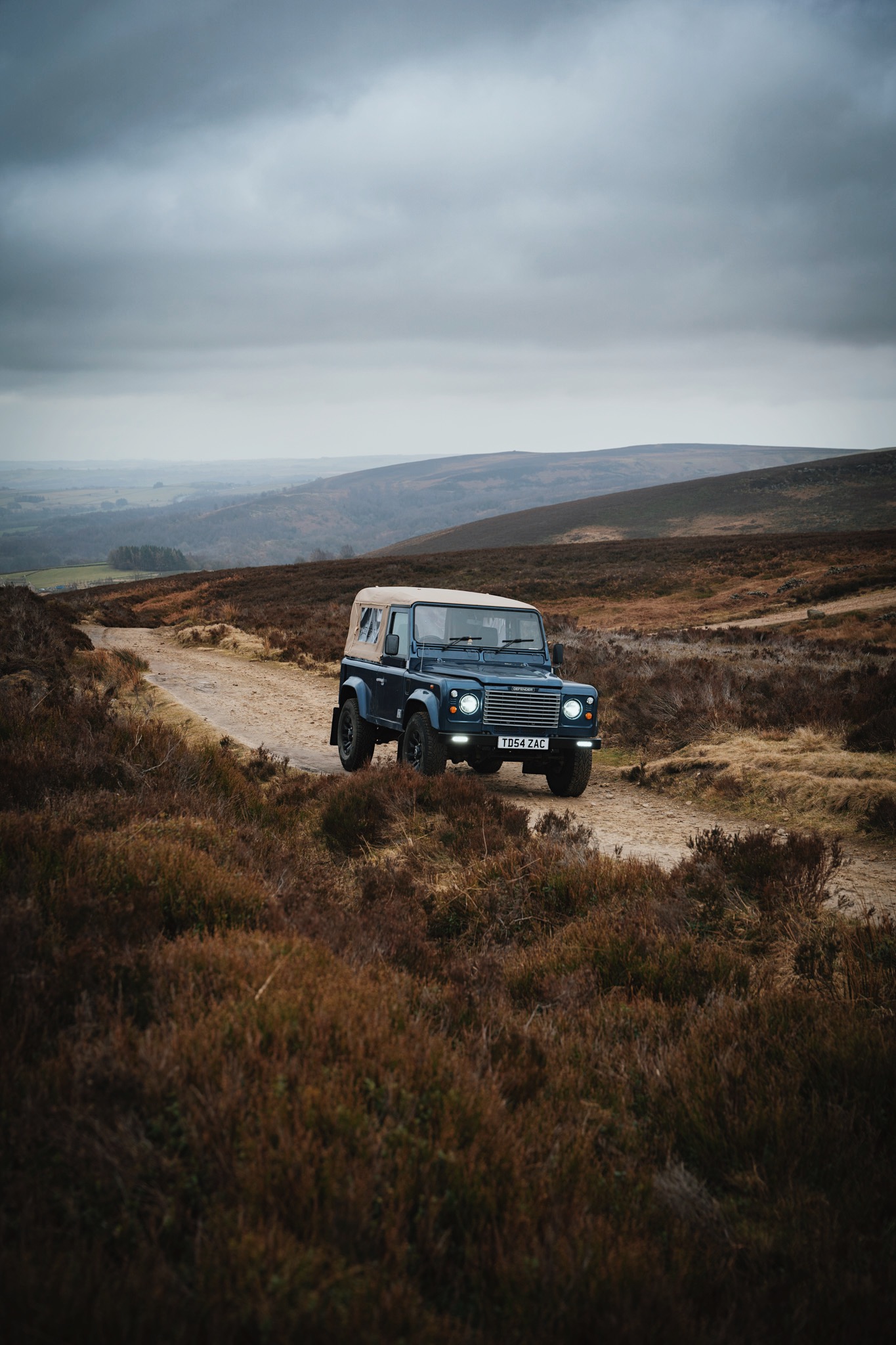 Blue off-road vehicle with beige roof driving on a dirt path through moorland under cloudy sky.