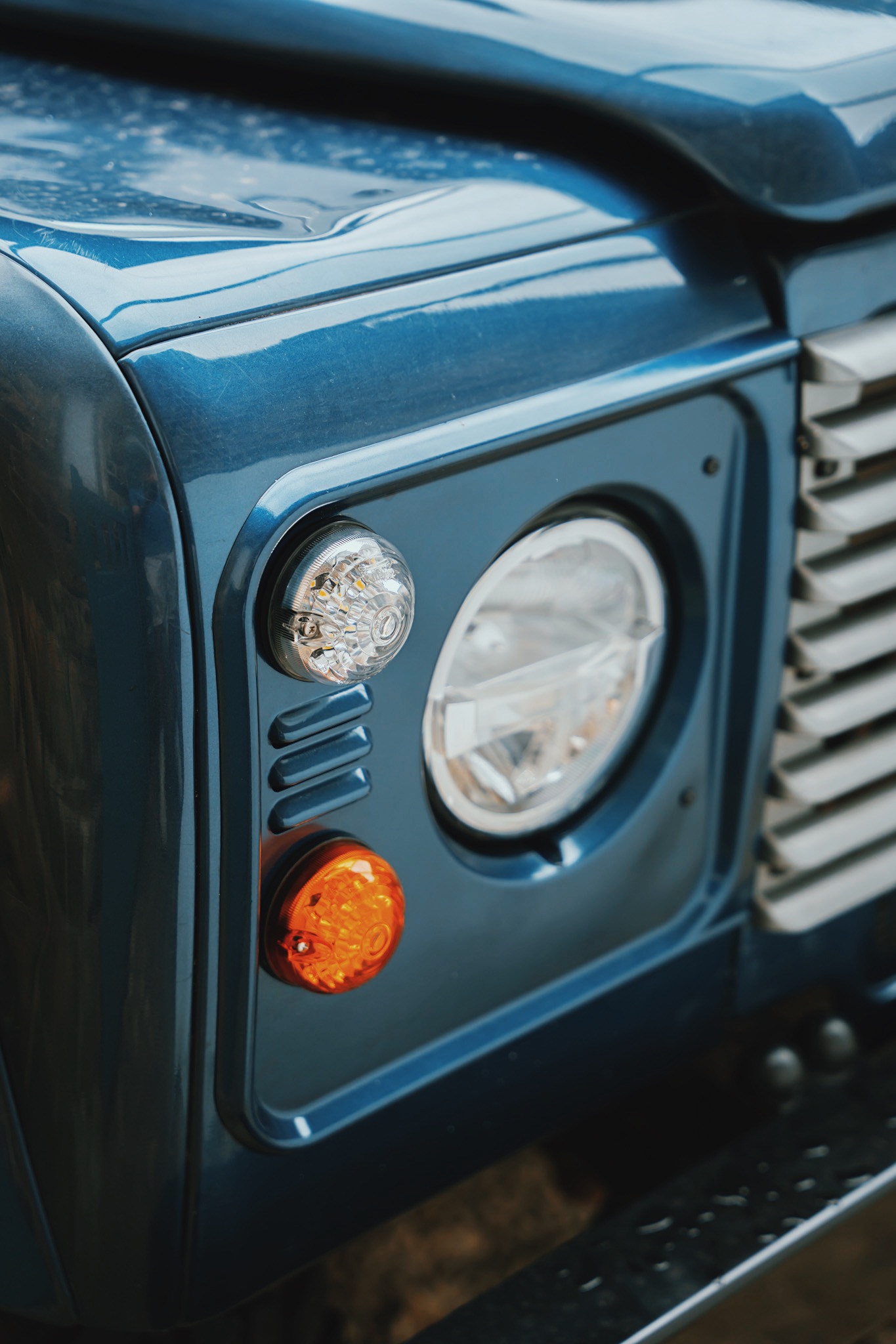 Close-up of the front left corner of a blue vehicle showing headlight, amber turn signal, and part of the grille.