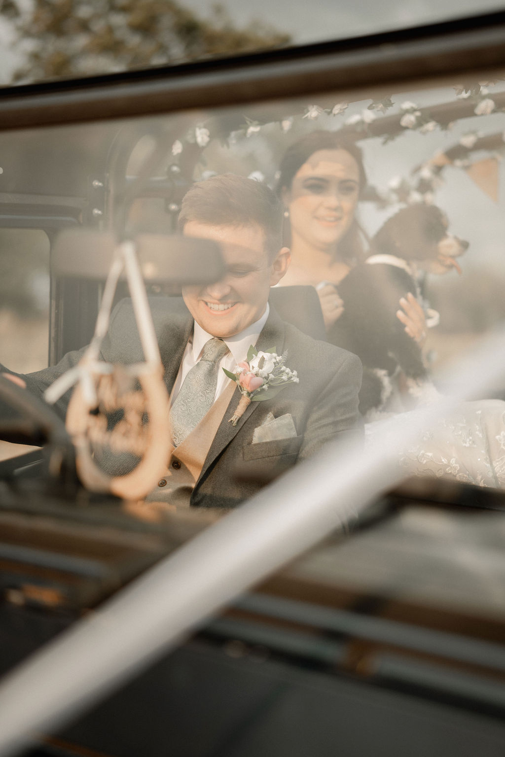 Smiling groom in suit sitting in a car with bride holding a small black and white dog in the back seat.