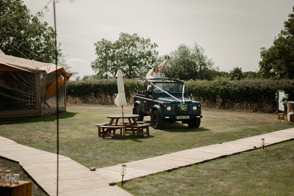 Bride in a white wedding dress standing in a decorated blue off-road vehicle holding up a bouquet on a grassy outdoor wedding venue.