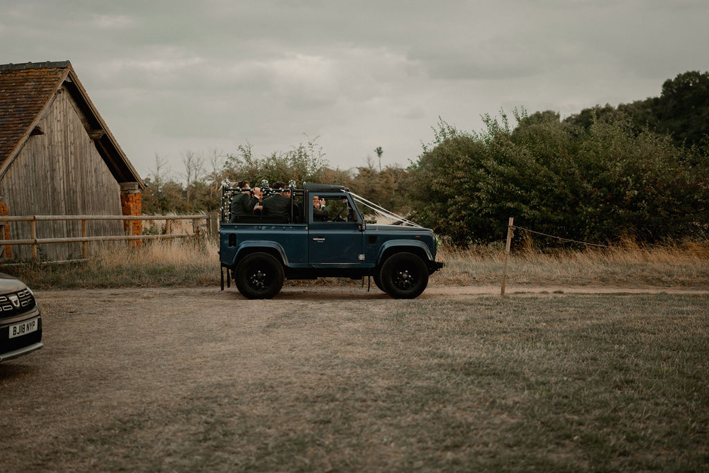 Blue open-top vehicle with people inside parked on a dirt path near a wooden fence and rustic building.