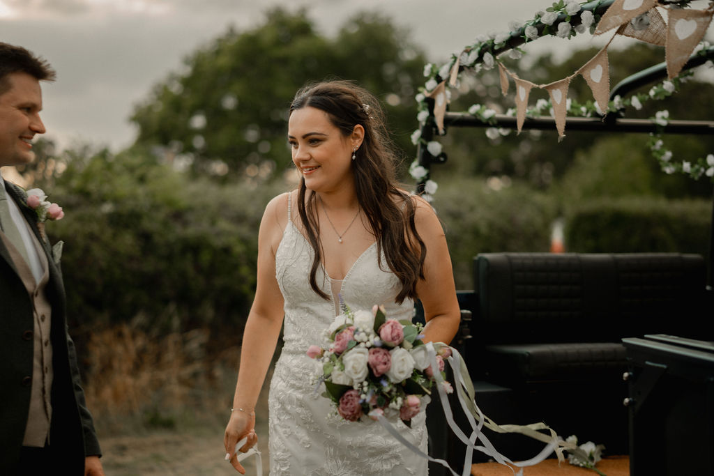 Bride in white wedding dress holding a bouquet of pink and white flowers next to a groom in outdoor setting.