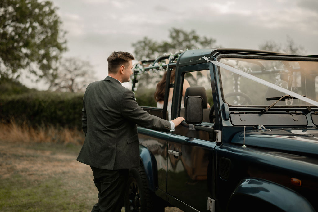 Man in a suit standing next to a decorated blue open-top vehicle with a woman seated inside.