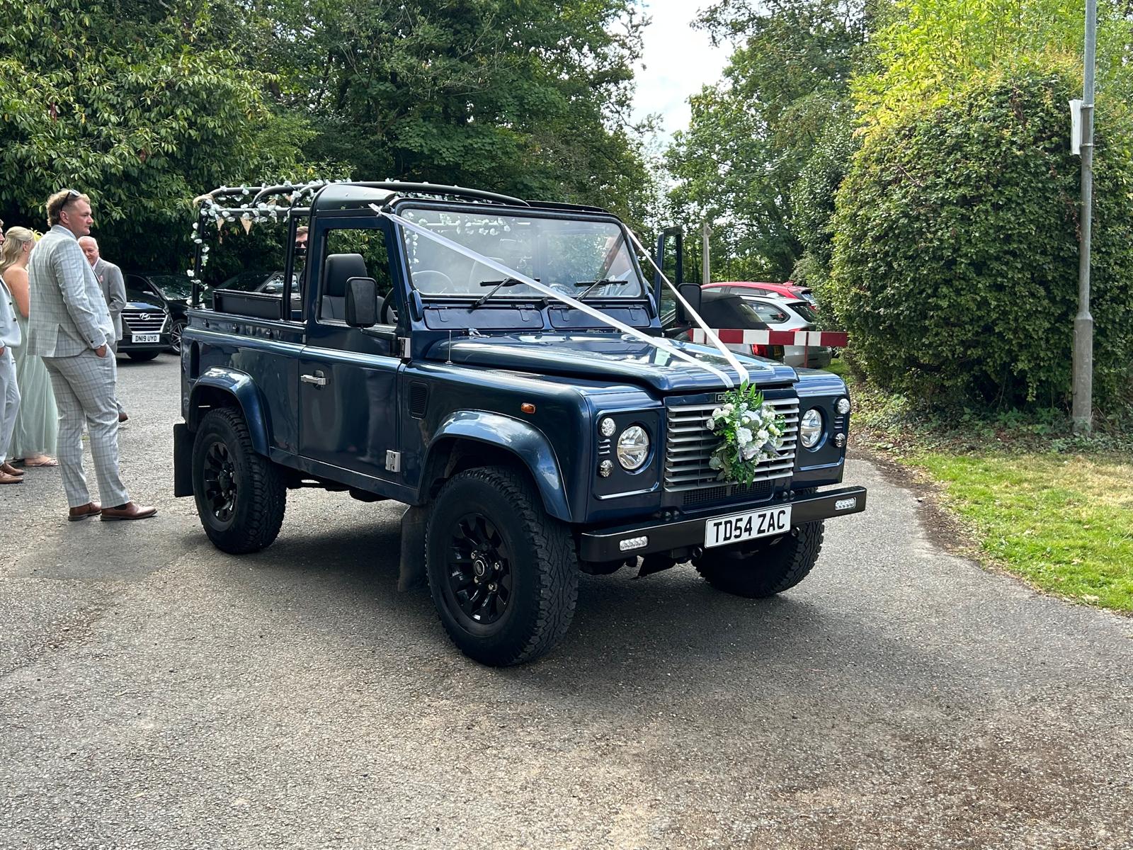 Dark blue Land Rover decorated with white flowers and ribbons parked on a road with people in formal attire nearby.