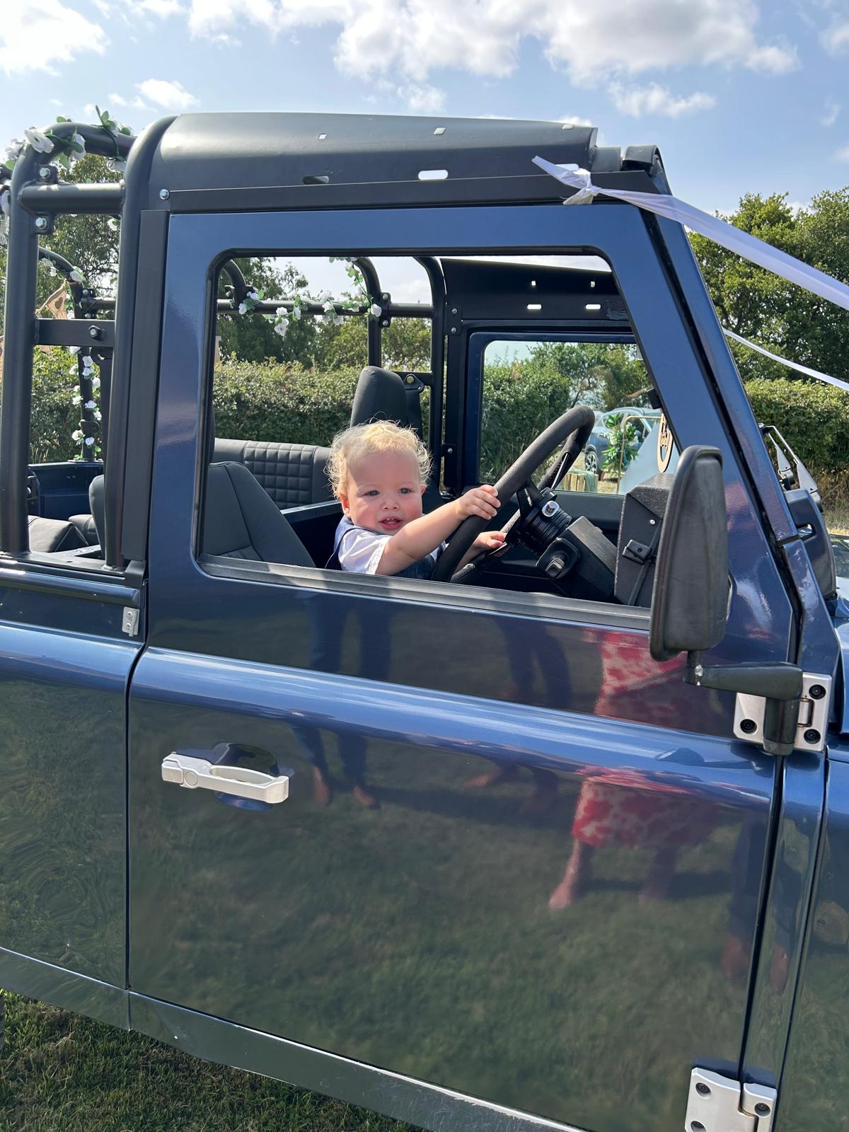 Young child sitting in the driver's seat of a blue open-top vehicle holding the steering wheel.