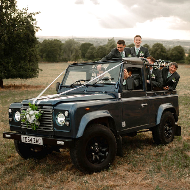 Four men in suits riding in a decorated dark blue off-road vehicle in a grassy field at sunset.