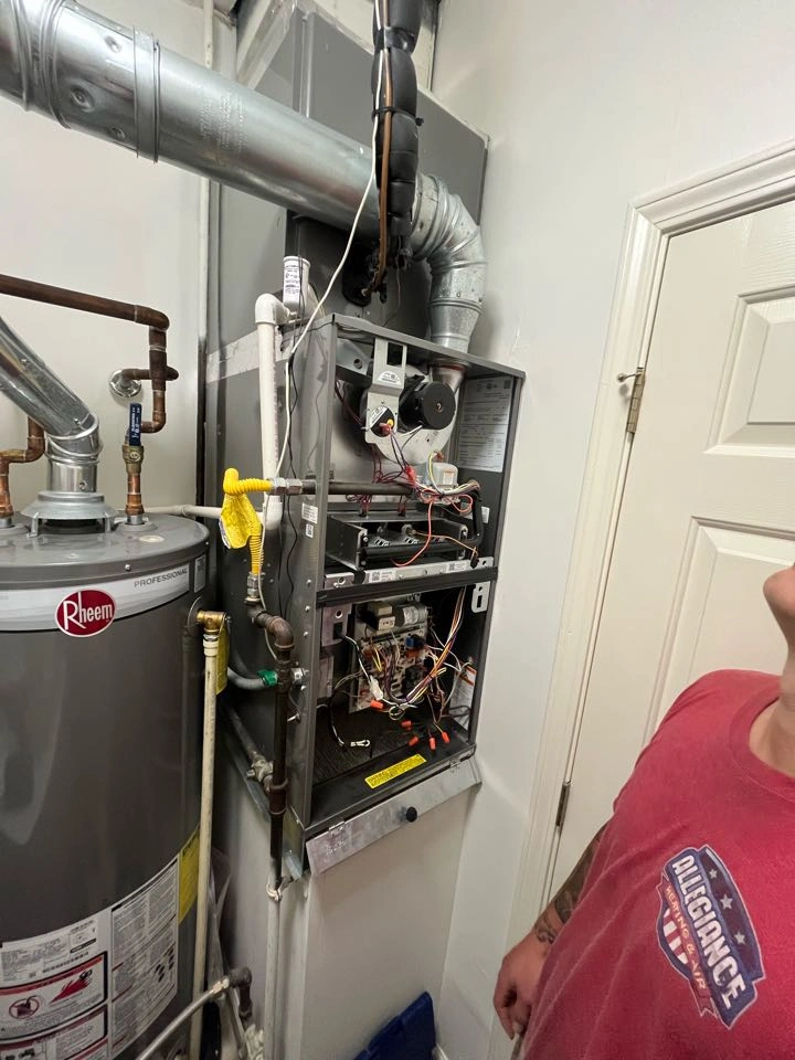 A man observes a furnace with its panel open, exposing wiring and components.