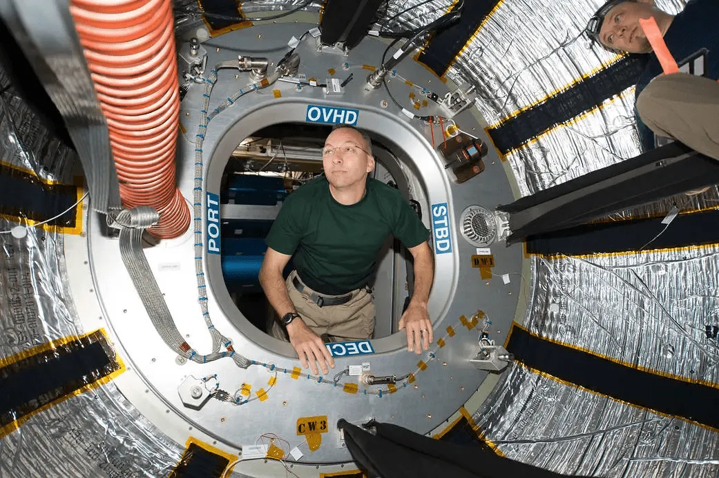 Two astronauts inside a space station module, one floating through a circular hatch labeled with 'OVHD', 'PORT', 'STBD', and 'DECI'.