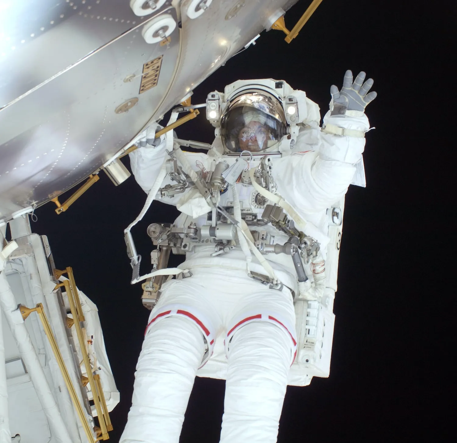 Astronaut in white suit waving while floating outside a spacecraft against the blackness of space.