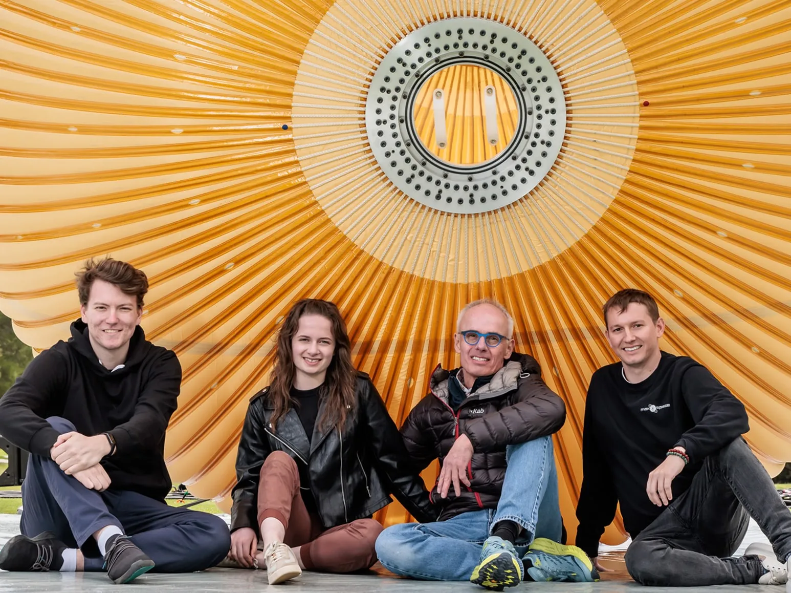 Four people sitting cross-legged smiling in front of a large yellow umbrella or canopy structure.
