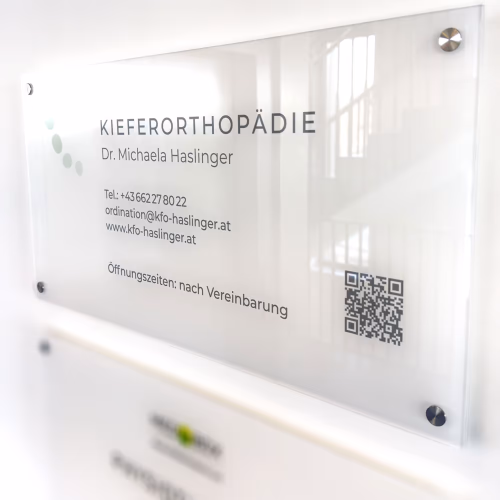 Schild an einer Wand mit der Aufschrift Kieferorthopädie Dr. Michaela Haslinger, Telefonnummer, Email, Webseite und QR-Code.