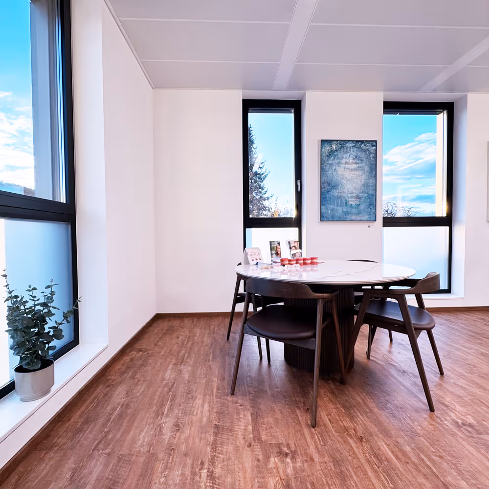 Moderner Besprechungsraum mit rundem Tisch, vier Stühlen, großen Fenstern und einem kleinen Topf mit Pflanze am Fenster.