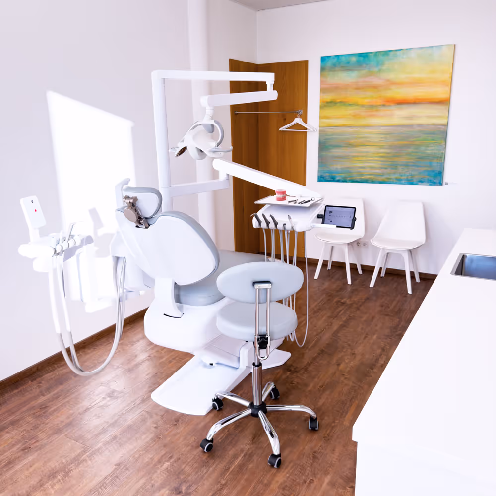 Helle Zahnarztpraxis mit Zahnarztstuhl, Stuhl auf Rollen, zwei weißen Stühlen an der Wand und einem bunten Bild.
