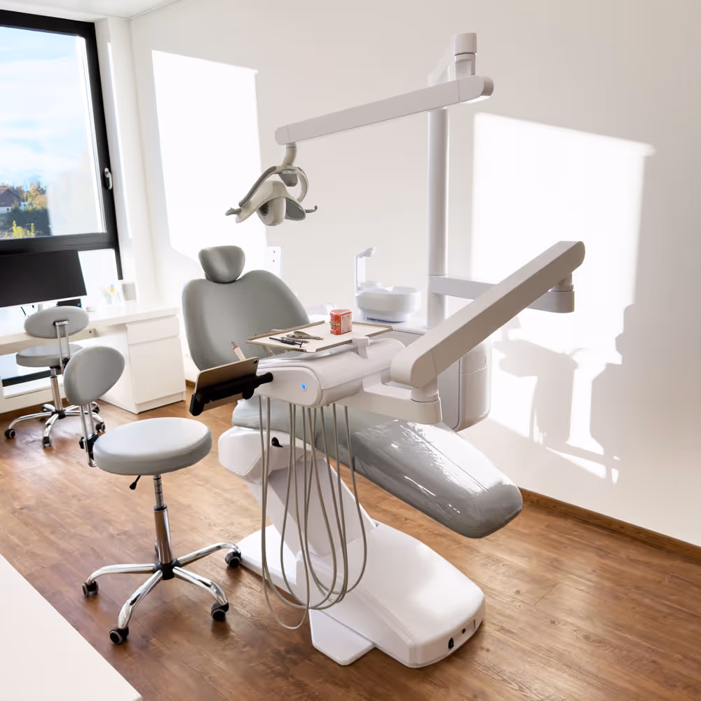 Heller Behandlungsraum mit weißem Zahnarztstuhl, Instrumenten und zwei weißen Hockern auf Holzfußboden.