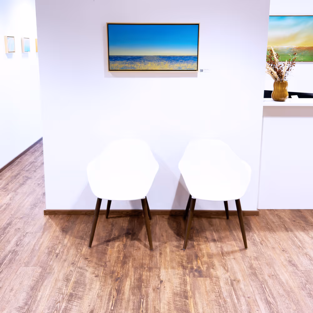 Zwei weiße Stühle vor einer weißen Wand mit einem Gemälde eines Meereslandschaft und Holzboden.