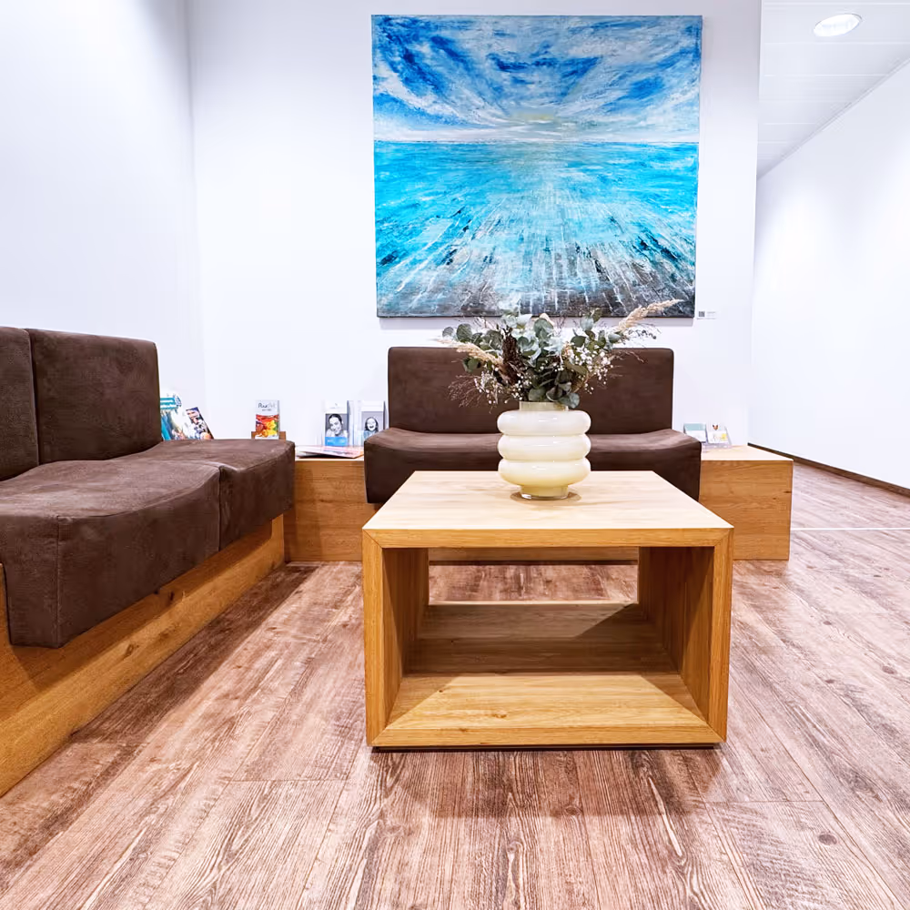 Warteraum mit braunen Sitzbänken, einem Holztisch mit Vase und abstraktem Blau-grauem Gemälde an der Wand.